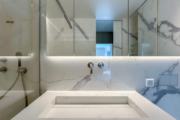 Lavabo moderne sur un comptoir avec carrelage imitation marbre Calacatta de Graniti Fiandre, avec robinetterie encastrée et miroir réfléchissant, dans une salle de bains à Nyon.